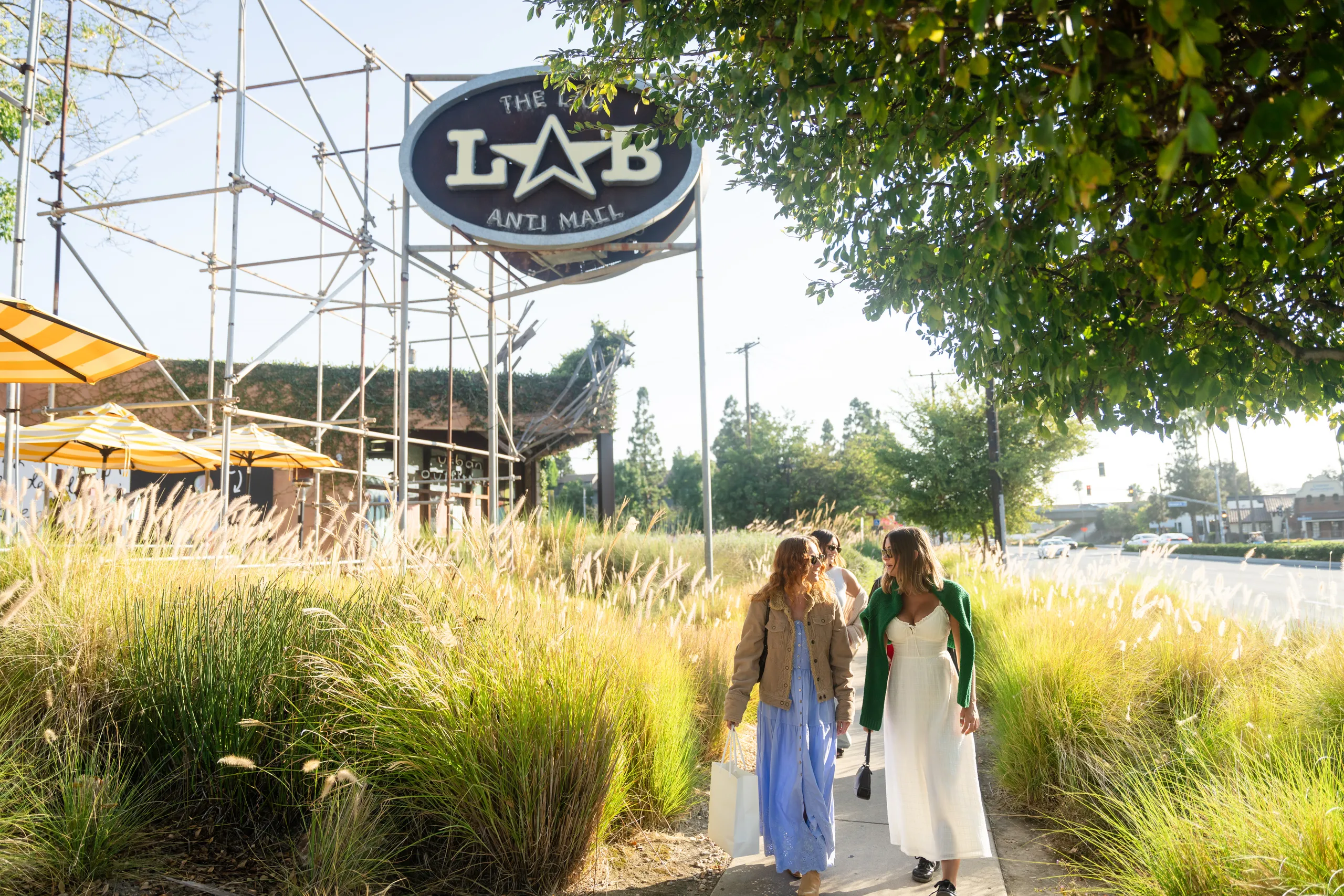 Three women walk along a sunlit sidewalk near tall grass at The Lab Costa Mesa, carrying shopping bags beneath a large sign that reads The Lab Anti-Mall, with trees and umbrellas in the background.