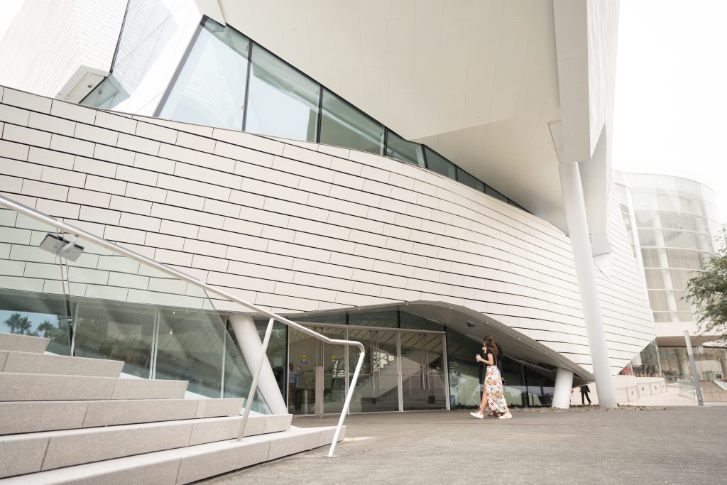 A woman in a long, floral skirt stands near the modern, angular exterior of a white building with large windows and tiled walls in Costa Mesa. A staircase with a metal railing leads up on the left side.