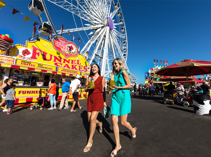 Two women holding drinks walk and laugh at a fair, with a large Ferris wheel, food stands including a funnel cake booth, and crowds of people enjoying the sunny day in the background.