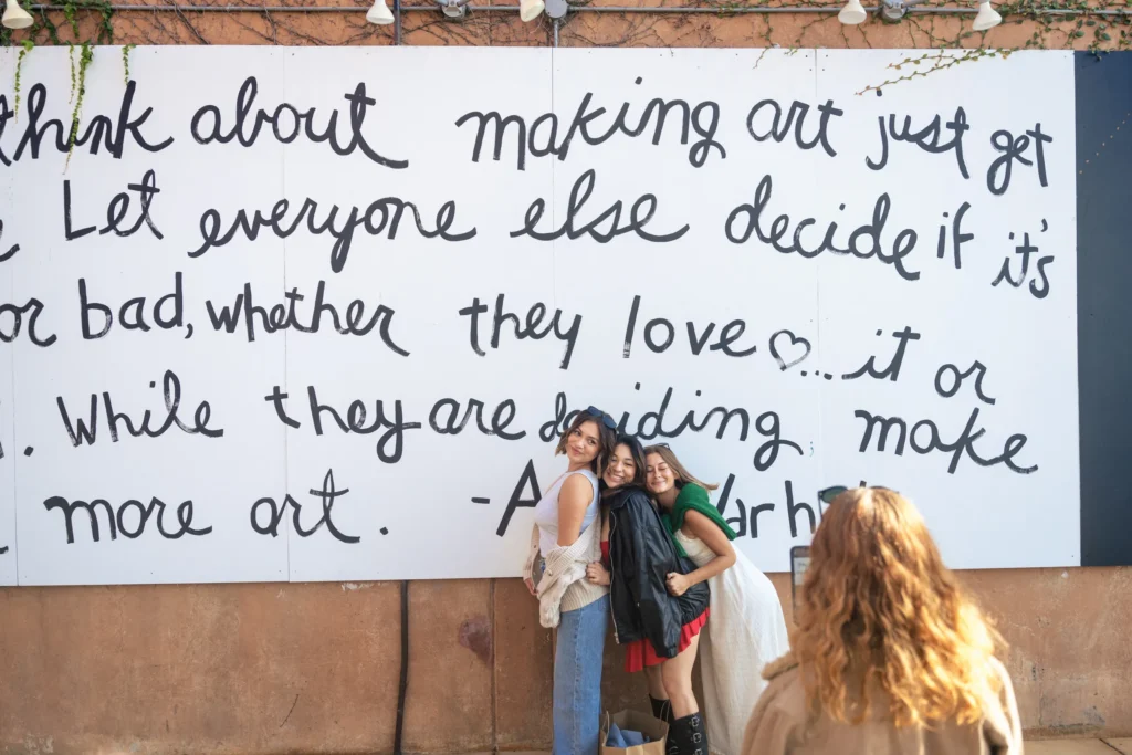 Three women pose and smile in front of a large mural with handwritten text about art, perfect for a Costa Mesa travel blog. Another person with curly hair faces them, possibly taking a photo. The mural is on a tan wall with greenery above.