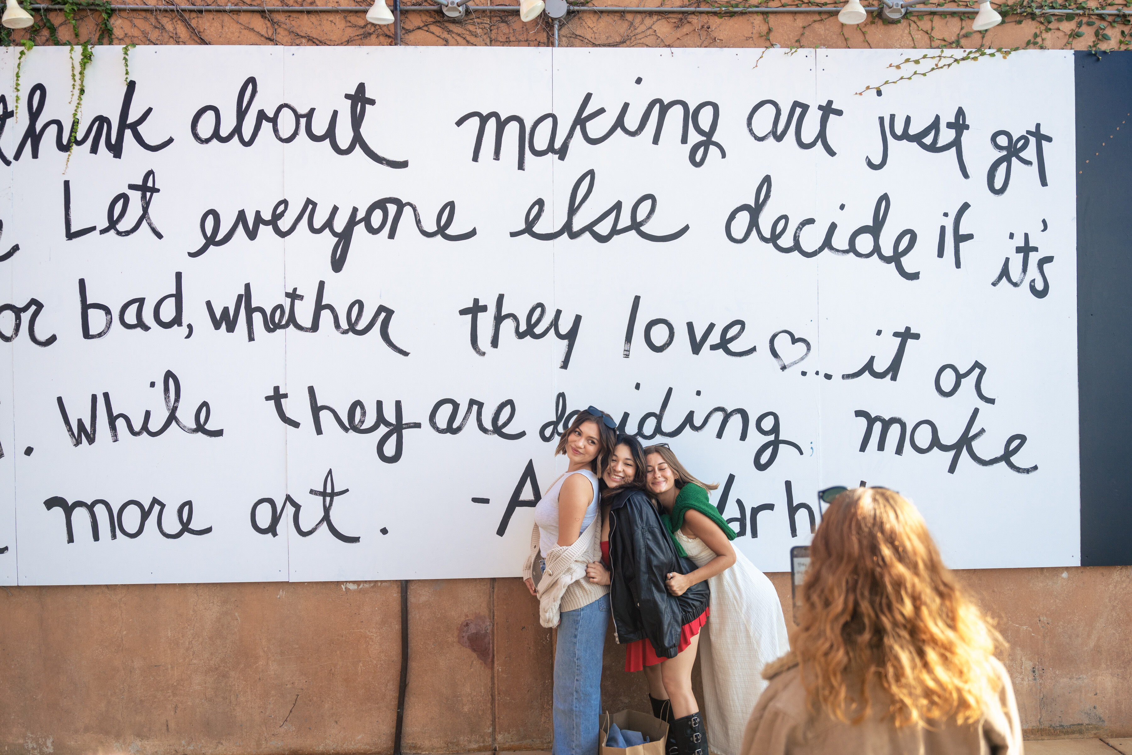Three women pose and smile in front of a large mural with handwritten text about art, perfect for a Costa Mesa travel blog. Another person with curly hair faces them, possibly taking a photo. The mural is on a tan wall with greenery above.