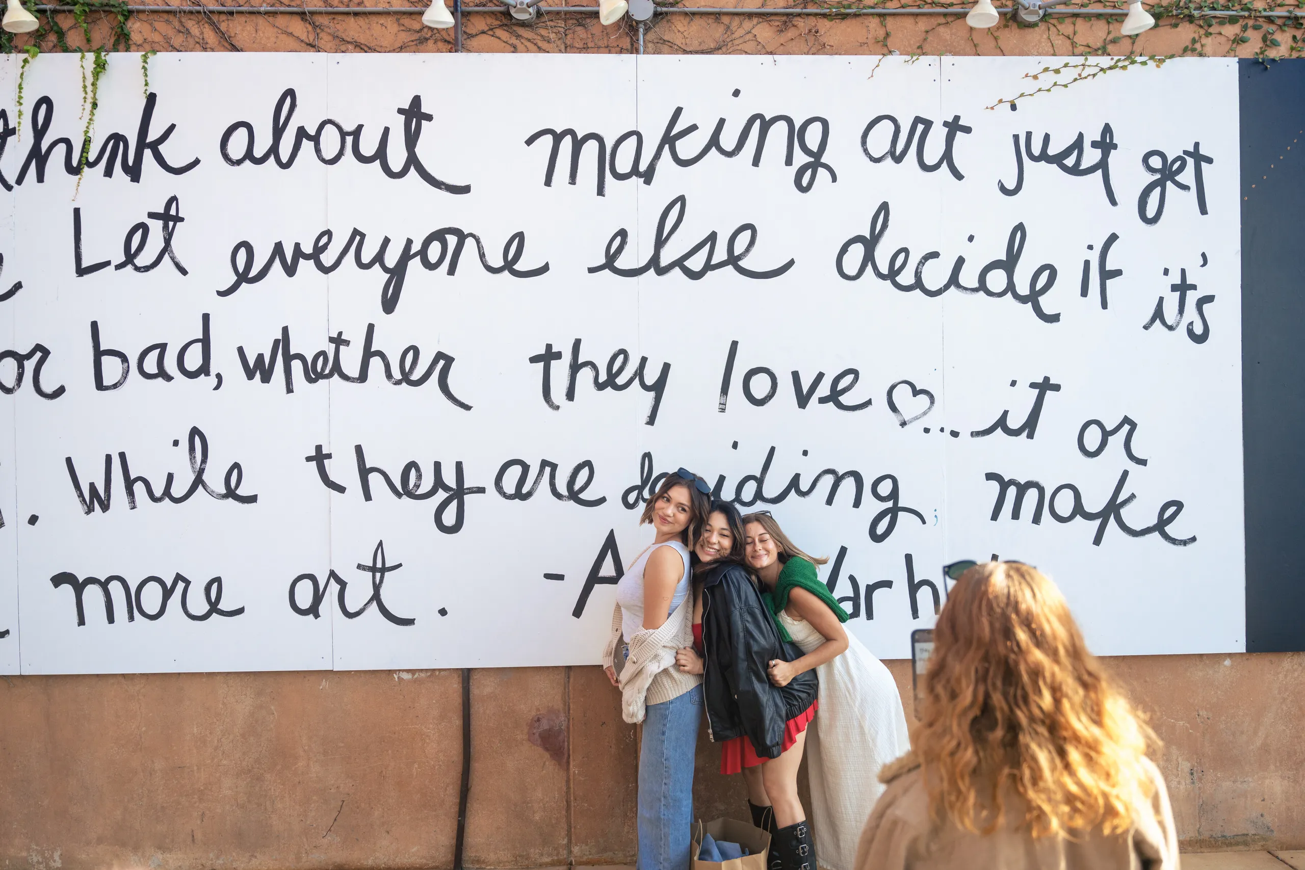 Three women pose and smile in front of a large mural with handwritten text about art, perfect for a Costa Mesa travel blog. Another person with curly hair faces them, possibly taking a photo. The mural is on a tan wall with greenery above.