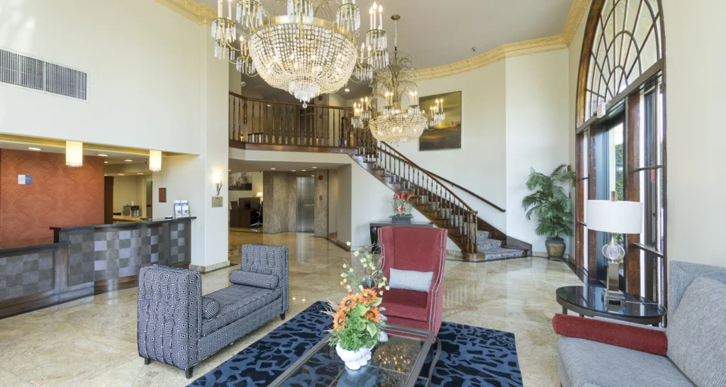Elegant hotel lobby with marble floors, large chandeliers, and a curved wooden staircase—an ideal setting for wedding venues Costa Mesa. Modern armchairs surround a glass coffee table topped with flowers, while large windows fill the space with natural light.