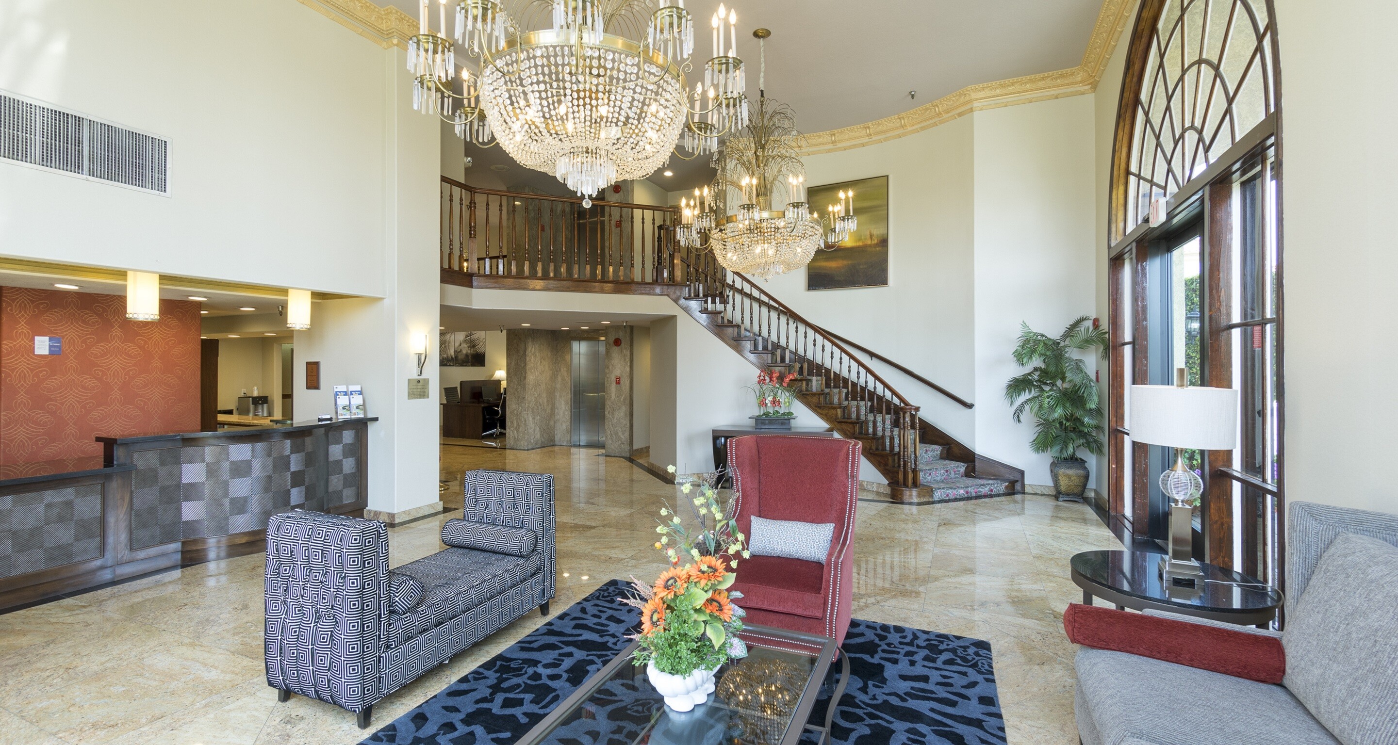 Elegant hotel lobby with marble floors, large chandeliers, and a curved wooden staircase—an ideal setting for wedding venues Costa Mesa. Modern armchairs surround a glass coffee table topped with flowers, while large windows fill the space with natural light.
