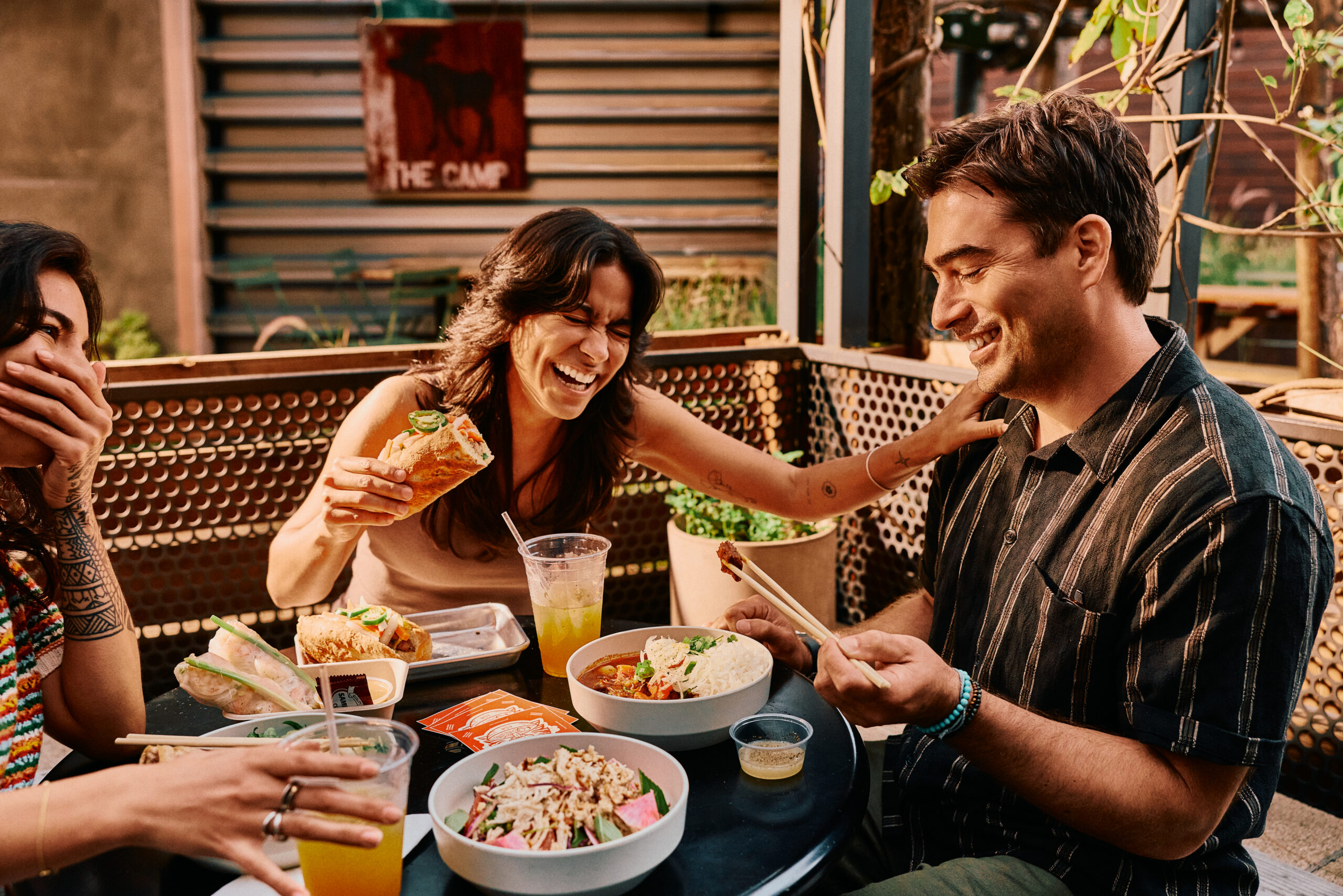 Three friends sit at an outdoor table in Costa Mesa, laughing and enjoying tacos, salads, and drinks. One person is holding a taco, while another checks their phone—the perfect eatcation vibe in a casual and cheerful setting.