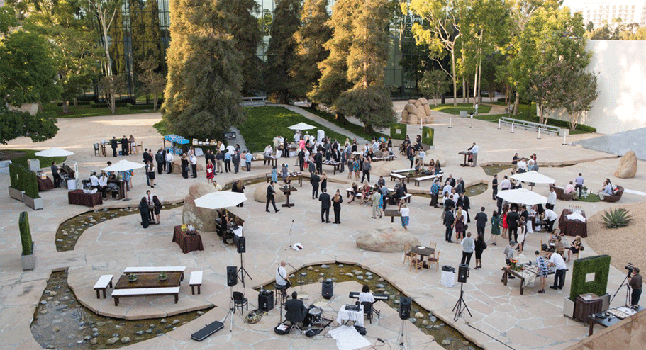 A large outdoor courtyard event at one of the top wedding venues Costa Mesa offers, with guests mingling, tables with white umbrellas, musical instruments set up, and modern landscaping featuring trees, rocks, and a small stream.