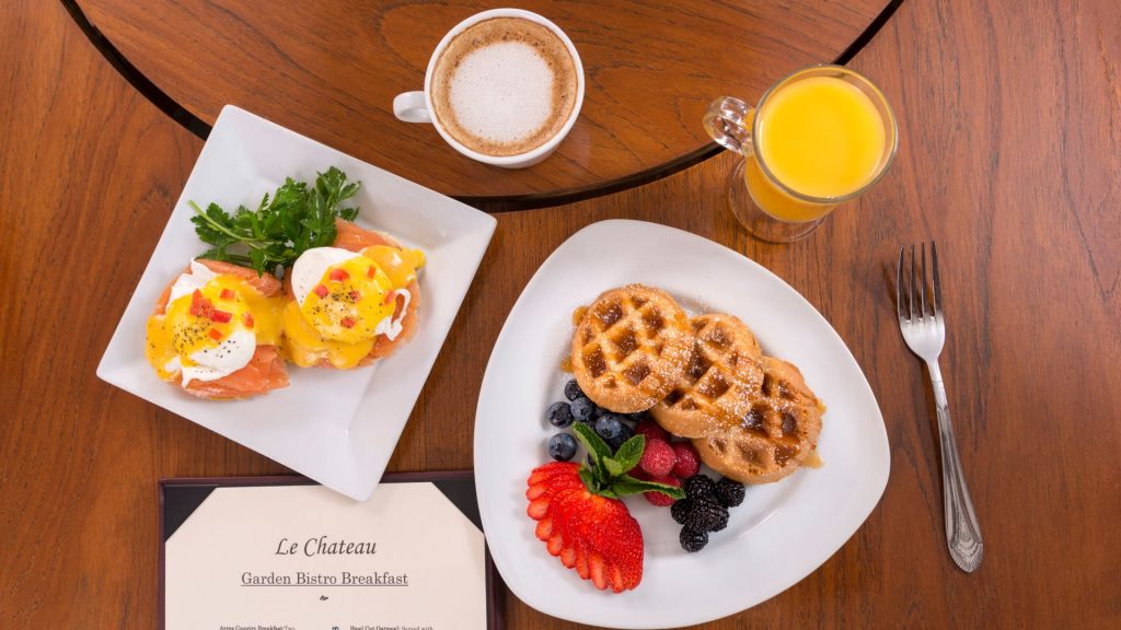 A breakfast spread with eggs benedict on toast, waffles topped with berries, a cup of cappuccino, a glass of orange juice, and a menu on a wooden table.