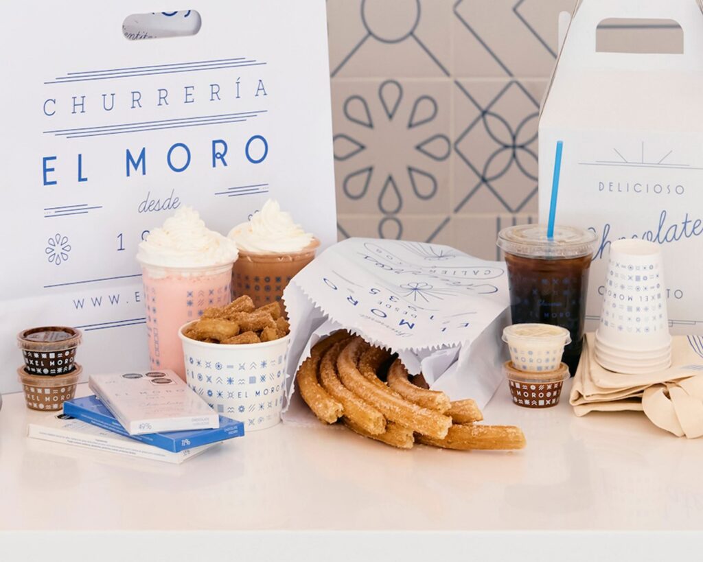 A variety of churros, dipping sauces, hot chocolate, coffee, and cold drinks on a white counter, with branded El Moro packaging—reminiscent of the minimalist blue-and-white decor found at Silver Trumpet Restaurant Costa Mesa.