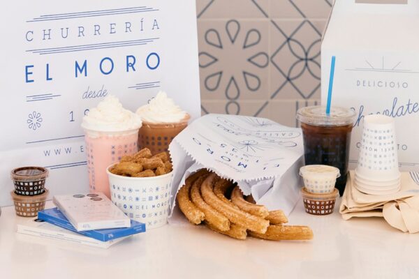 A variety of churros, dipping sauces, hot chocolate, coffee, and cold drinks on a white counter, with branded El Moro packaging—reminiscent of the minimalist blue-and-white decor found at Silver Trumpet Restaurant Costa Mesa.