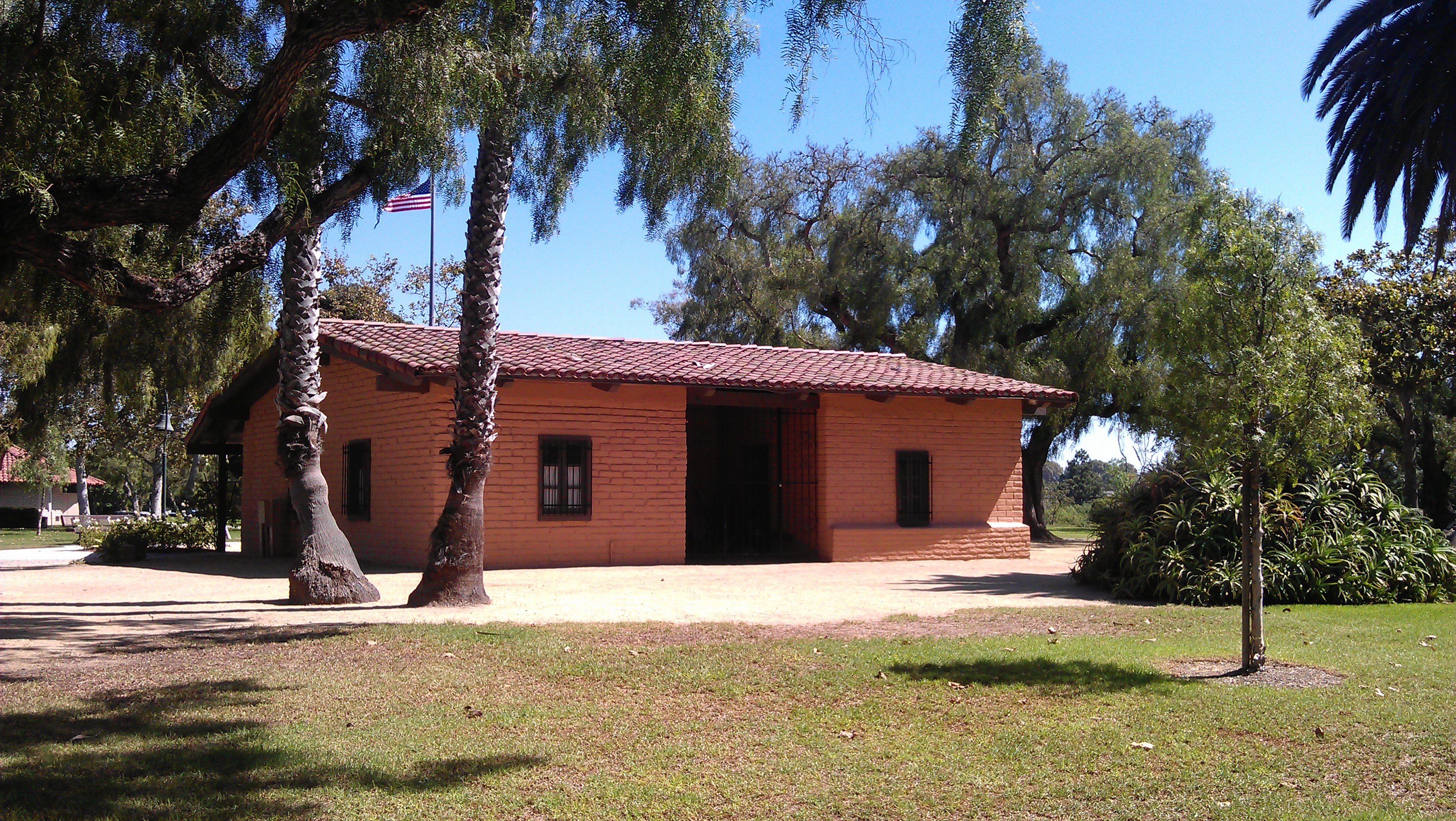 A small adobe building with a red tile roof sits in Estancia Park, Costa Mesa, surrounded by palm trees and green grass under a clear blue sky. An American flag is visible in the background.