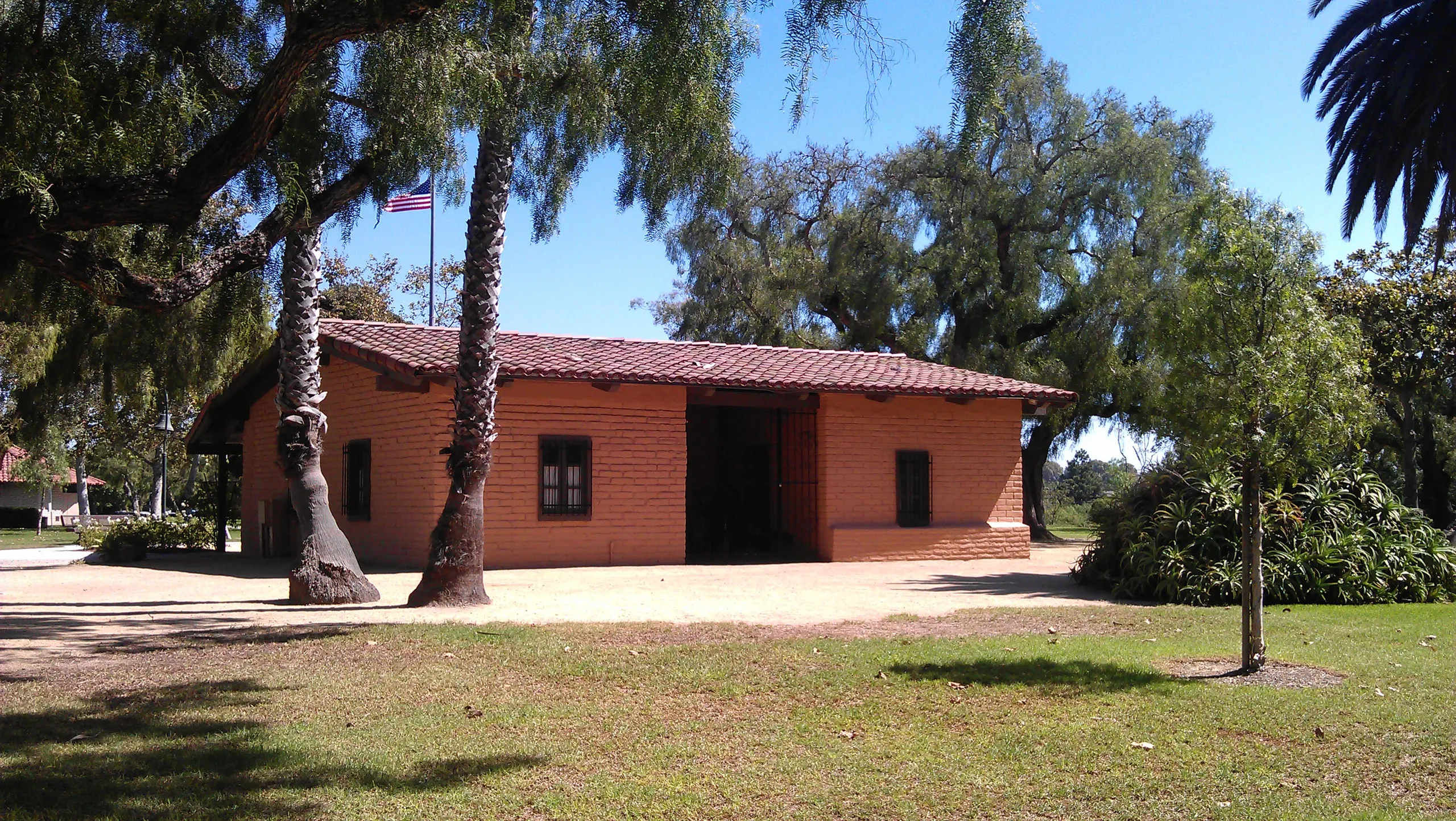 A small adobe building with a red tile roof sits in Estancia Park, Costa Mesa, surrounded by palm trees and green grass under a clear blue sky. An American flag is visible in the background.