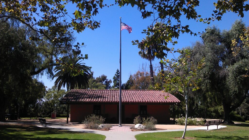 A brick building with a red-tiled roof sits behind a tall flagpole flying the American flag at Estancia Park Costa Mesa, surrounded by trees and benches on a sunny day.