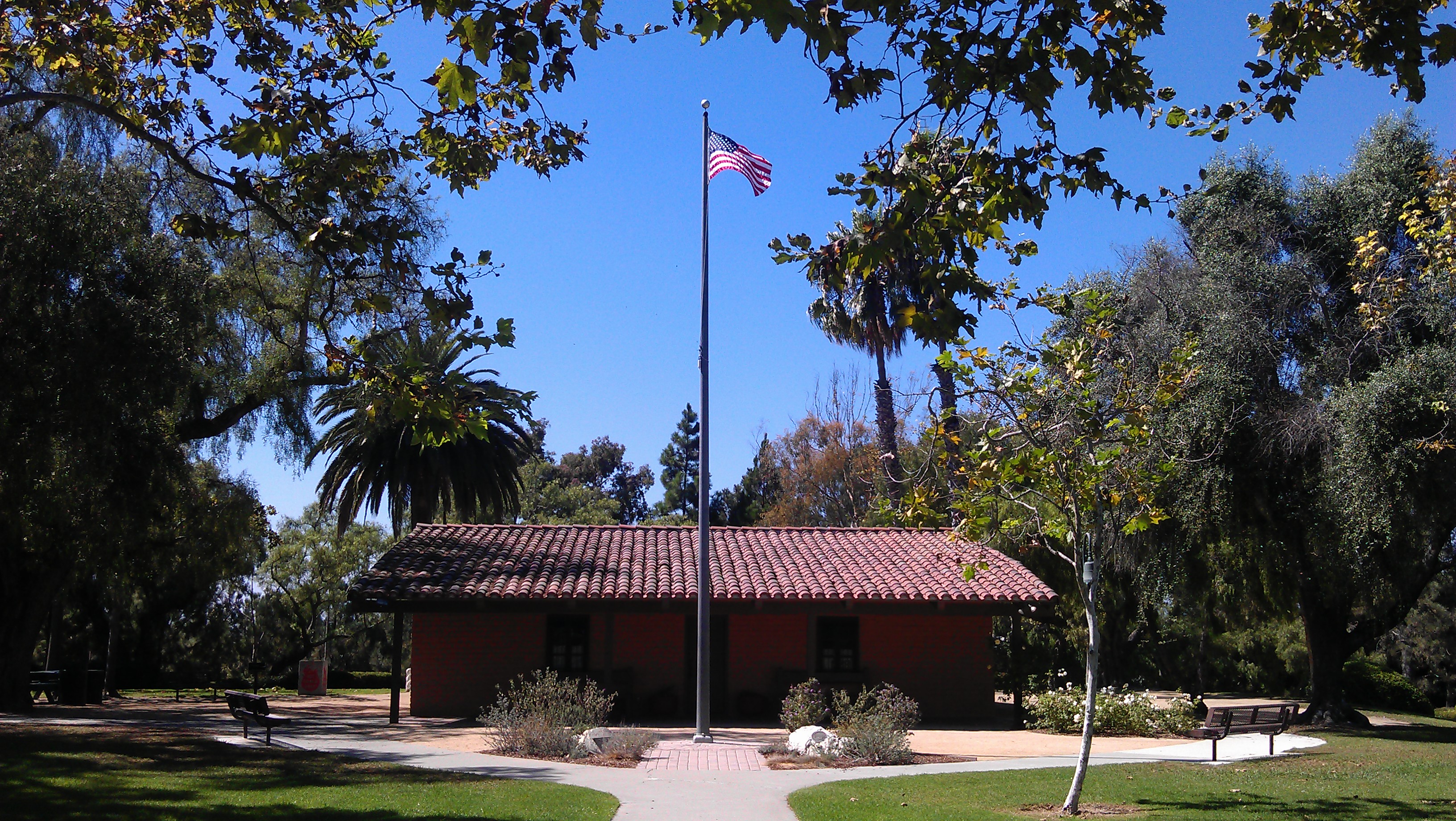 A brick building with a red-tiled roof sits behind a tall flagpole flying the American flag at Estancia Park Costa Mesa, surrounded by trees and benches on a sunny day.