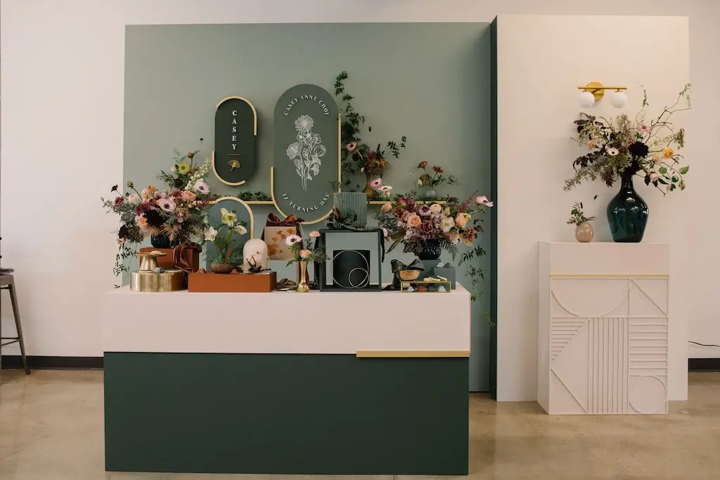 A modern display features floral arrangements, decorative tableware, and kitchen items set against green and white panels—perfect inspiration for a Costa Mesa travel blog, with framed botanical art and a vase with flowers on a side pedestal.