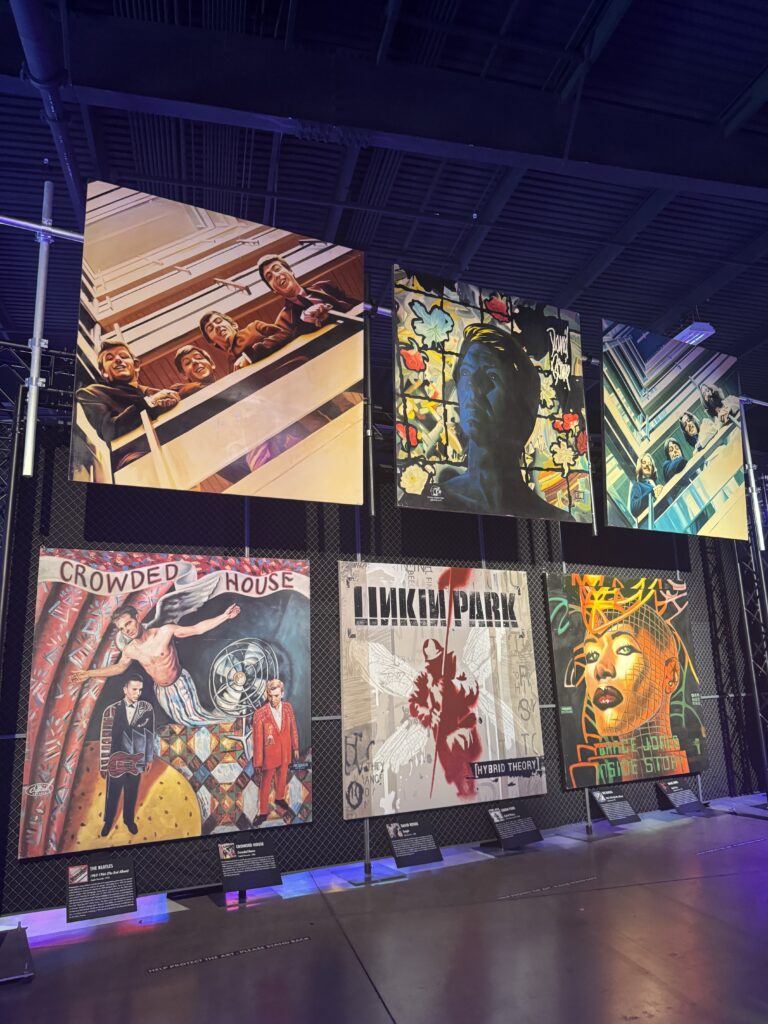 Six large, colorful album cover artworks are displayed in two rows at an indoor exhibit at the OC Fair Costa Mesa, featuring bands like Crowded House and Linkin Park, with spotlights illuminating the vibrant designs.