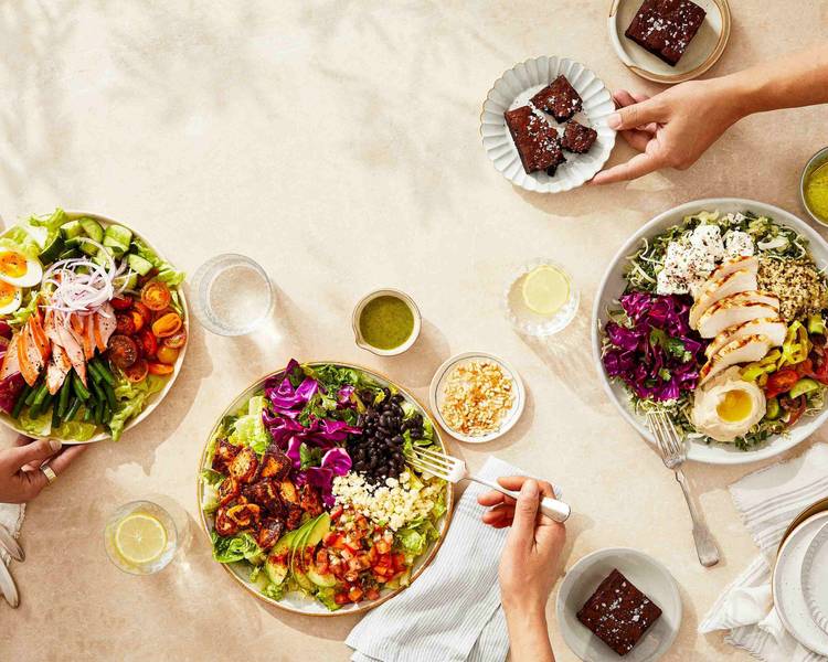 Three people are enjoying fresh salads topped with ingredients like grilled chicken, vegetables, avocado, black beans, and grains. There are brownies, dressings, lemon water, and small bowls of toppings on a light tabletop.