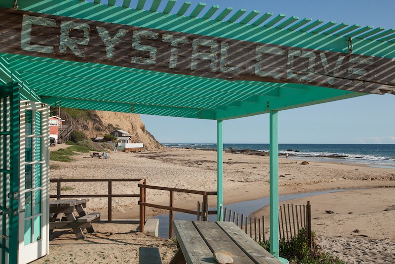 A turquoise pergola with CRYSTAL COVE written on top shades picnic tables on a sandy beach near Costa Mesa, with ocean waves, a wooden fence, and cliffs in the background under a clear blue sky.