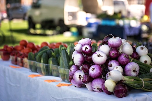 A table at an outdoor farmers’ market displays fresh produce, including piles of cucumbers, tomatoes, and a large stack of purple and white onions, with blurred greenery and vehicles in the background.