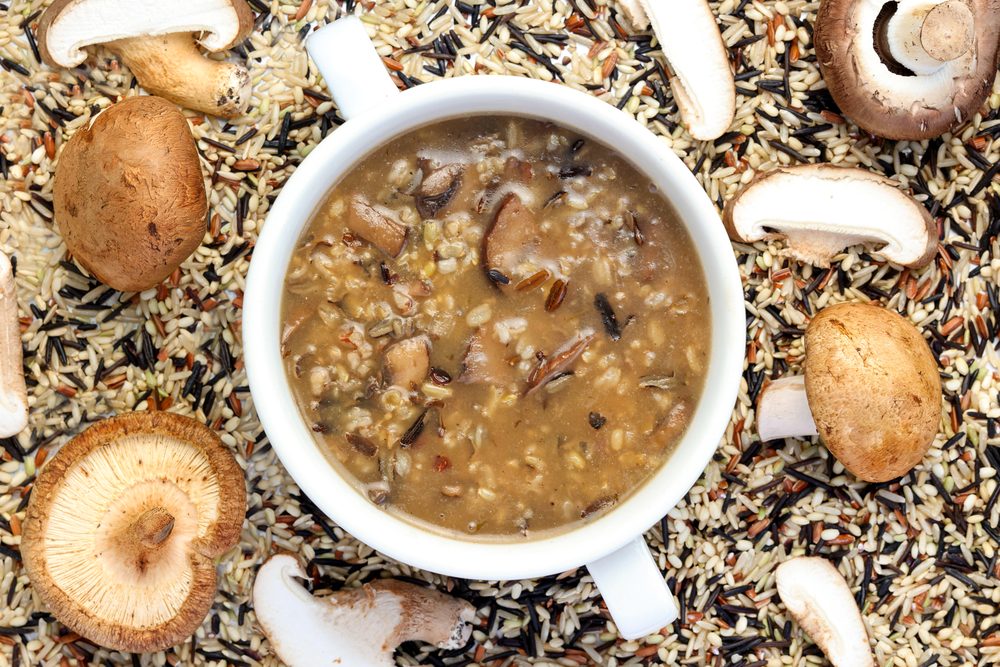 A bowl of mushroom and wild rice soup sits on a surface covered with uncooked wild rice and assorted whole and sliced mushrooms. The soup appears hearty with visible mushroom pieces and rice.