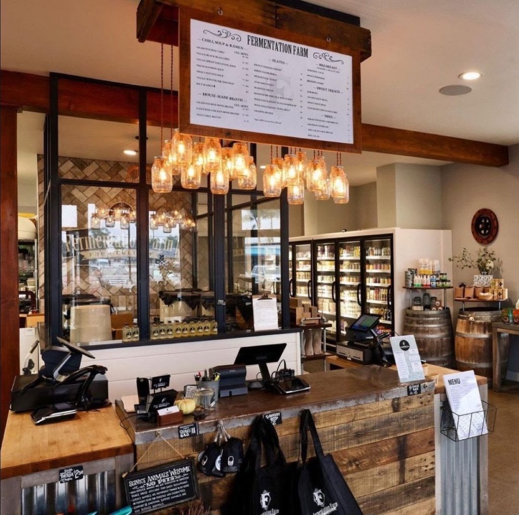 Rustic store counter with hanging jar lights, wooden surfaces, cash registers, black shopping bags, a large menu sign above, and refrigerators with various products in the background. Decor is warm and inviting.