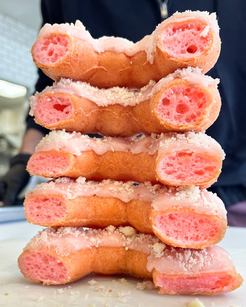 A stack of pink, frosted donuts with a bubbly texture, neatly arranged in a vertical pile. The donuts are cut in half, revealing their airy interior and topped with a light sprinkle of crumbs.