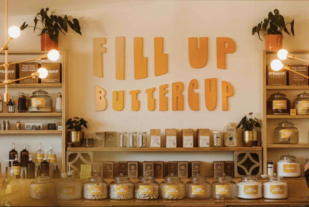 A shop interior with shelves holding large glass jars and boxes, featuring a prominent yellow sign on the wall that reads FILL UP BUTTERCUP in playful letters.