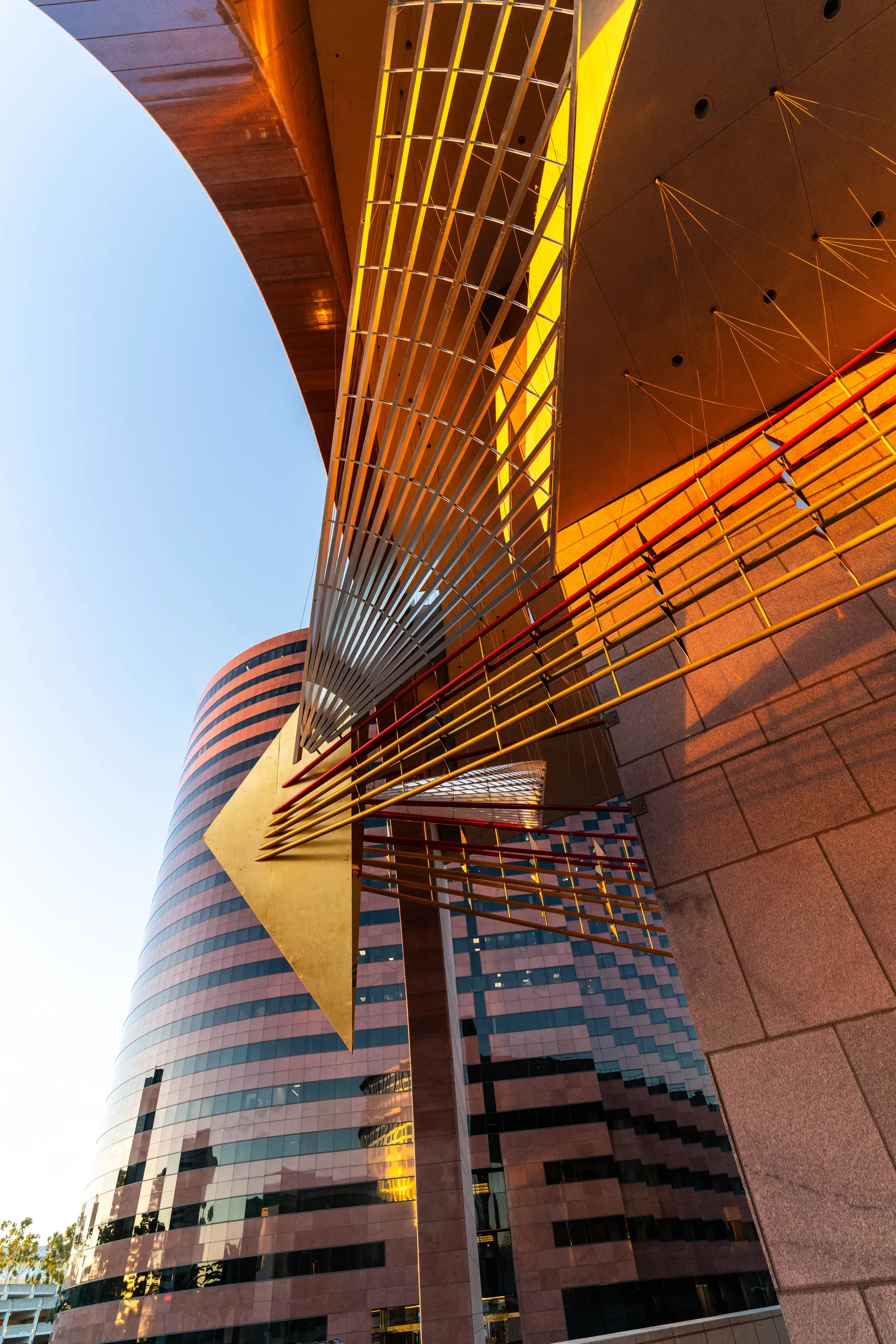 Modern architectural detail with a geometric golden metal structure inspired by the Fire Bird Sculpture, suspended above a curved glass office building with pink and black stripes under a clear blue sky in Costa Mesa.