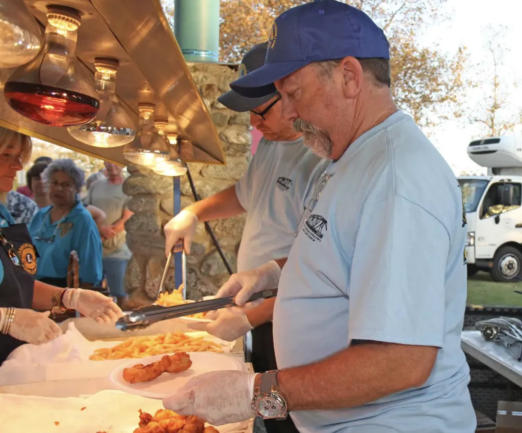 Two men in blue shirts and caps serve fried food and french fries onto plates at an outdoor food stand, while others wait in line. The servers wear gloves and work under a heat lamp. A truck is visible in the background.