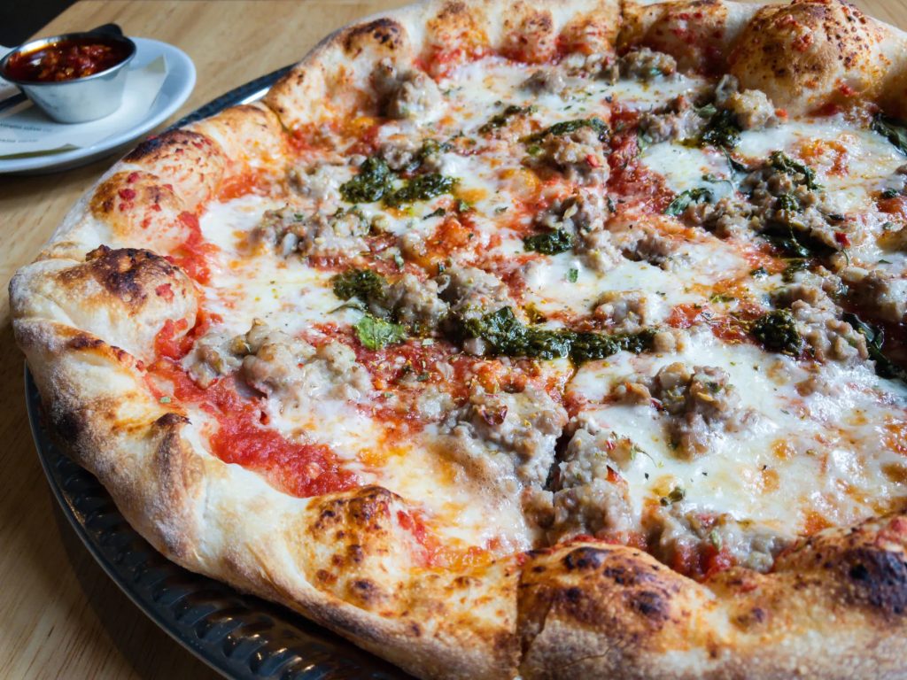 A close-up of a freshly baked pizza with a thick, golden crust, topped with melted cheese, crumbled sausage, tomato sauce, and herbs; a small dish of red sauce sits in the background.
