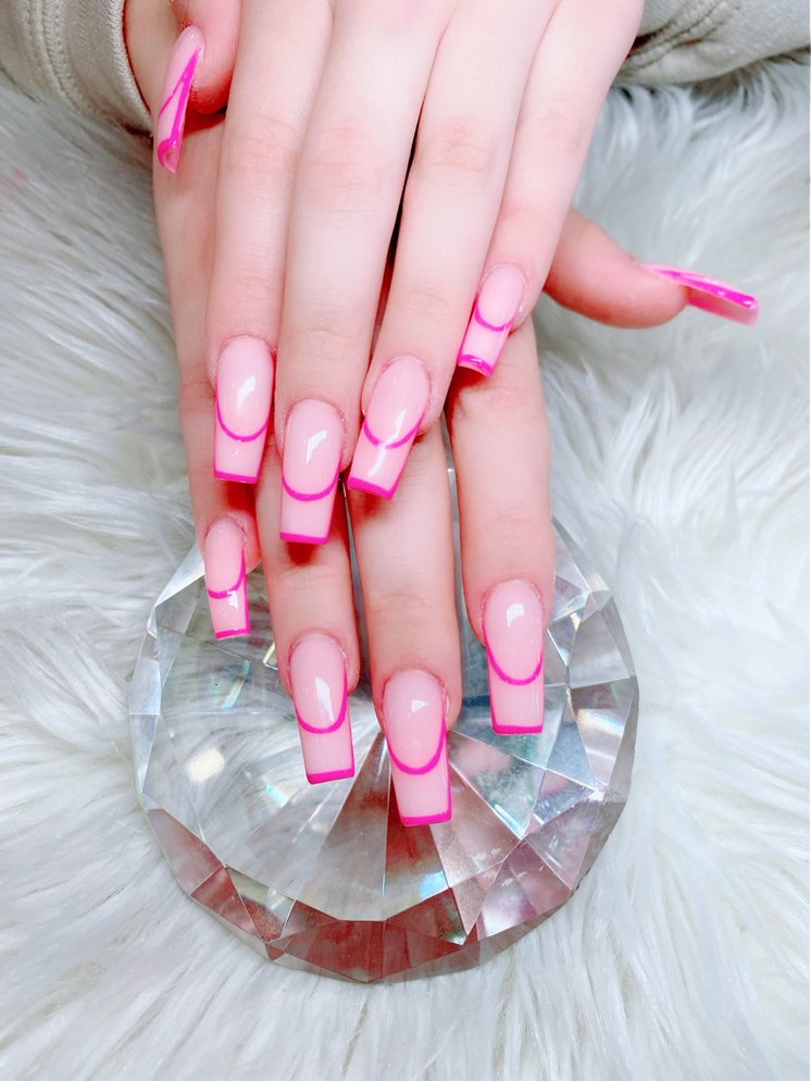 Two hands with long square acrylic nails featuring a light pink base and bright pink French tips are displayed over a large, faceted crystal on a white furry surface.