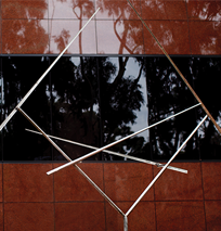 Thin metal rods intersect in an abstract geometric sculpture, featured along the Costa Mesa Art Walk, set against a reddish-brown tiled wall with a reflective black window showing tree silhouettes.