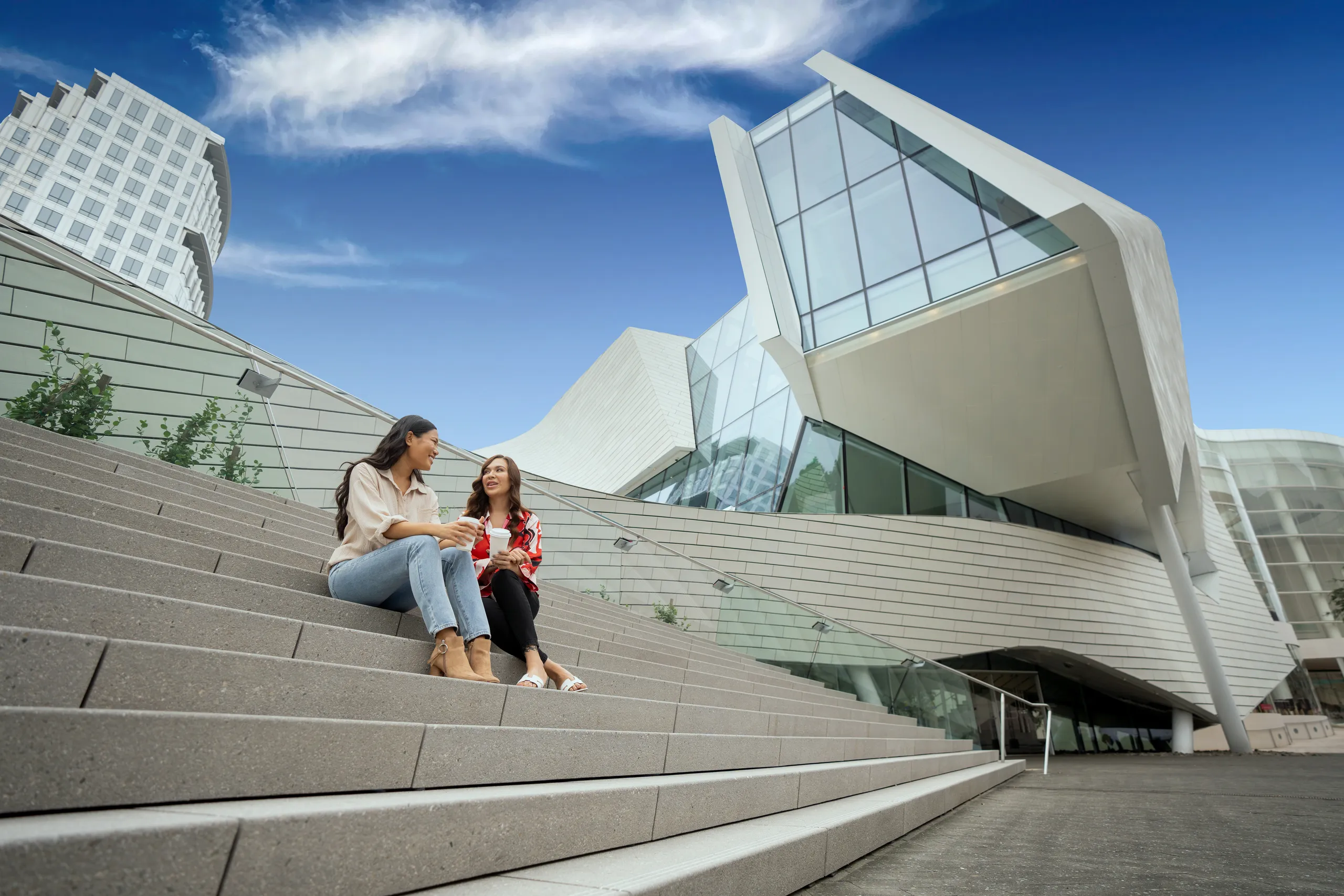 Two women sit on outdoor steps, chatting near the Orange County Museum of Art—a modern, angular building with large windows and a unique architectural design—under a bright blue sky with wispy clouds.
