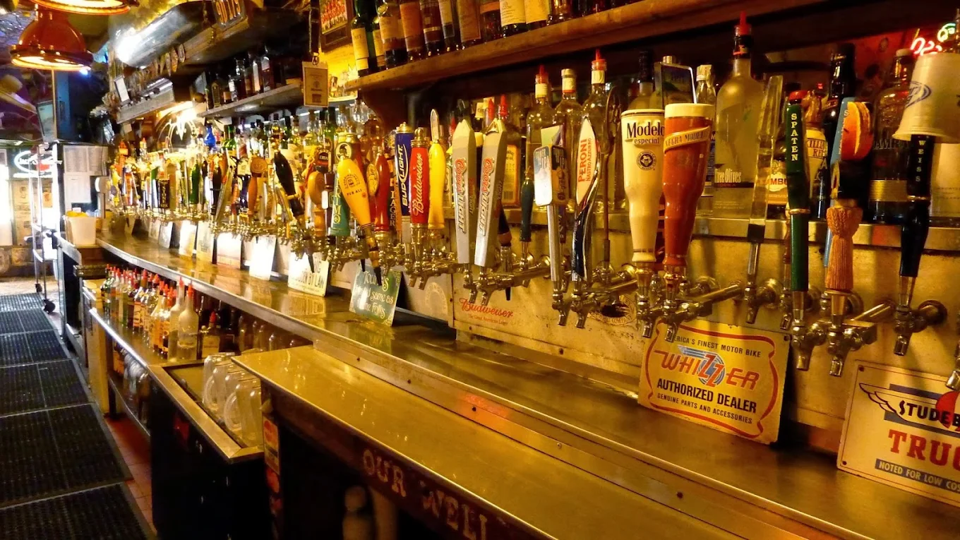 A long bar counter with dozens of beer taps featuring colorful handles at Goat Hill Tavern Costa Mesa, bottles of liquor on shelves above and below, and signs advertising different beer brands. The bar has a busy, lively atmosphere.