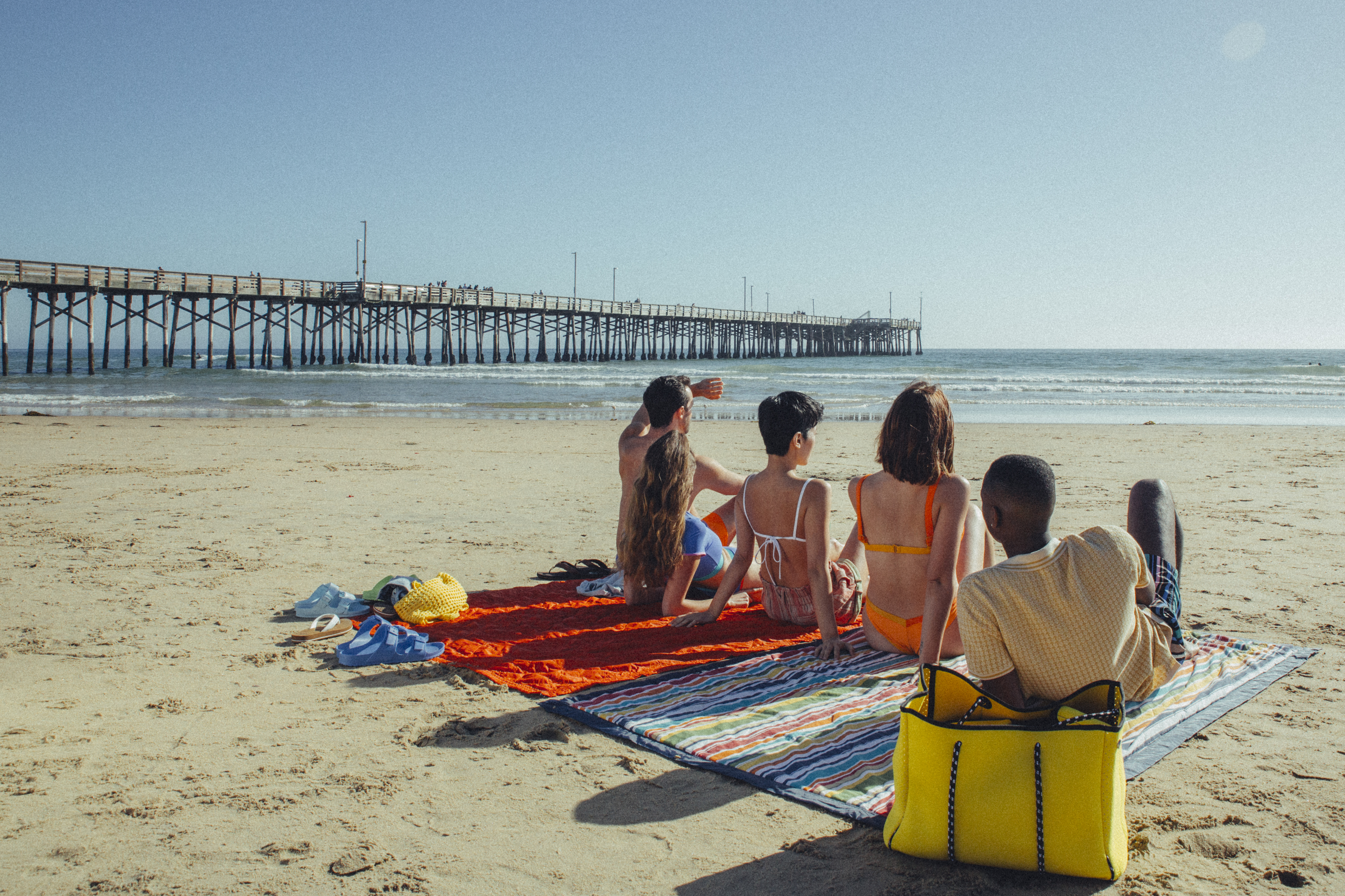 Five people sit on beach blankets facing the ocean at Newport Beach, with a wooden pier stretching into the water. The clear sky and various beach items, including a yellow bag, add to this classic Orange County scene.