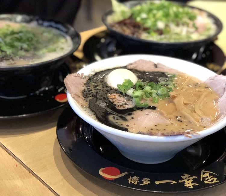 Three bowls of ramen on a table, with the closest bowl topped with sliced pork, green onions, a boiled egg, bamboo shoots, and a swirl of black garlic oil. The other two bowls are slightly blurred in the background.