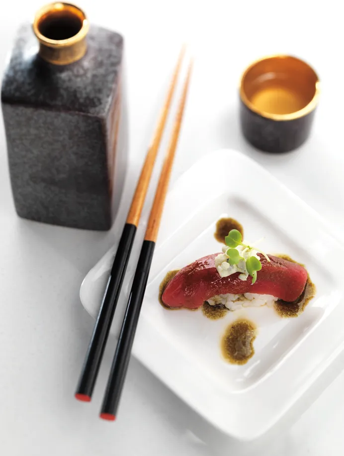 A single piece of Hamamori Costa Mesa tuna nigiri topped with sprouts on a small white plate, accompanied by chopsticks, a sake bottle, and a sake cup.