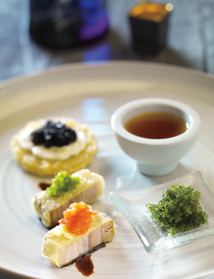 A plate of Hamamori Costa Mesa appetizers featuring a small tart with caviar, two pieces of fried fish topped with garnishes, a small bowl of dipping sauce, and a side of sea grapes.