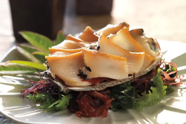 Slices of abalone served on a shell atop a bed of seaweed and greens at Hamamori Costa Mesa, with two dark square cups in the background.