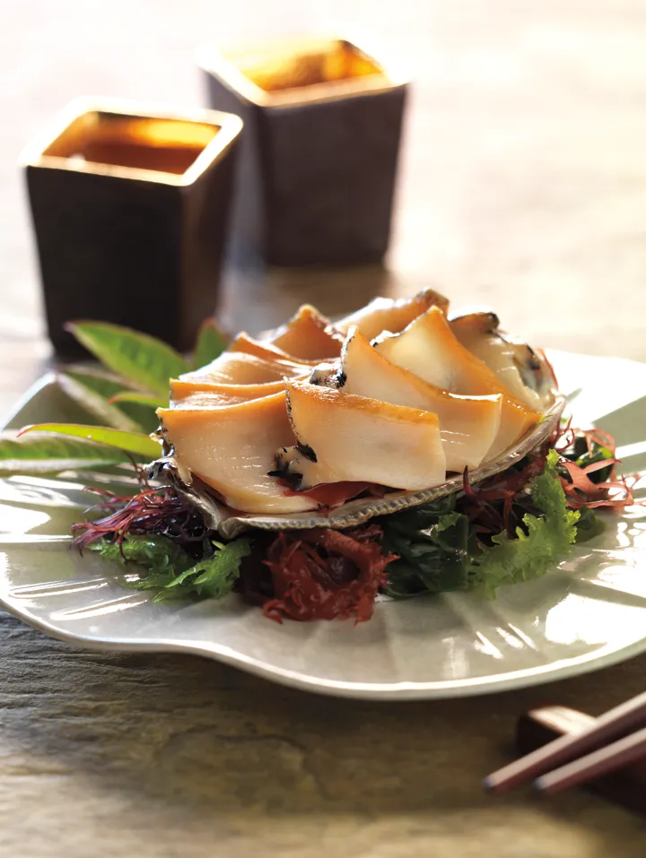 Slices of abalone served on a shell atop a bed of seaweed and greens at Hamamori Costa Mesa, with two dark square cups in the background.