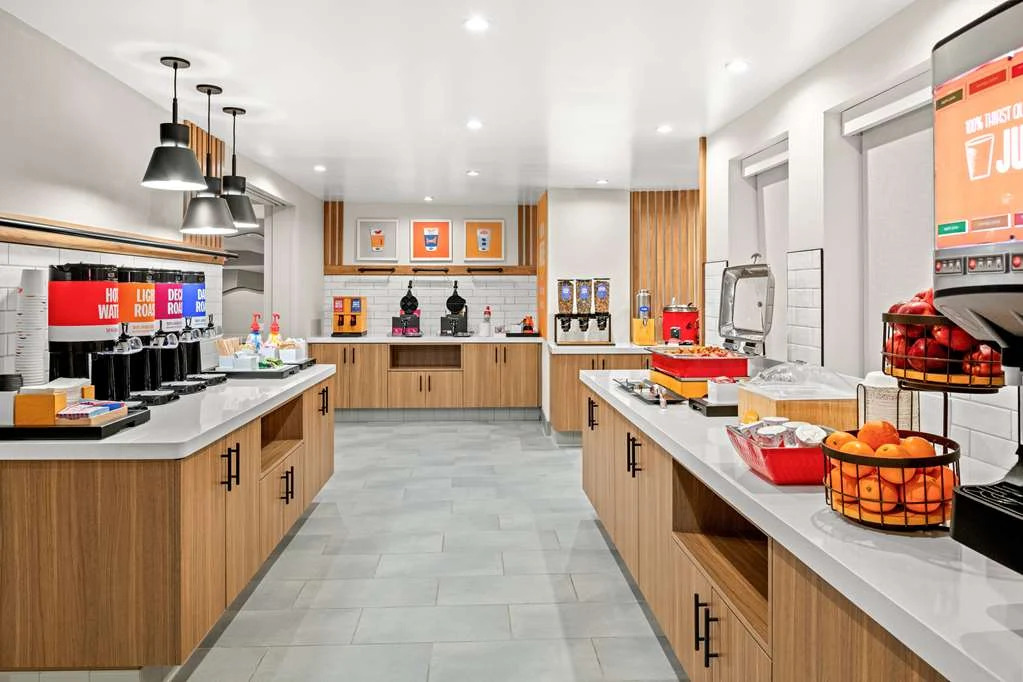 Hotel breakfast buffet area at Hampton Inn Costa Mesa Newport Beach features modern wood cabinets, white countertops, coffee and juice dispensers, cereal, pastries, and fresh fruit in a clean, inviting atmosphere with bright lighting and organized stations.