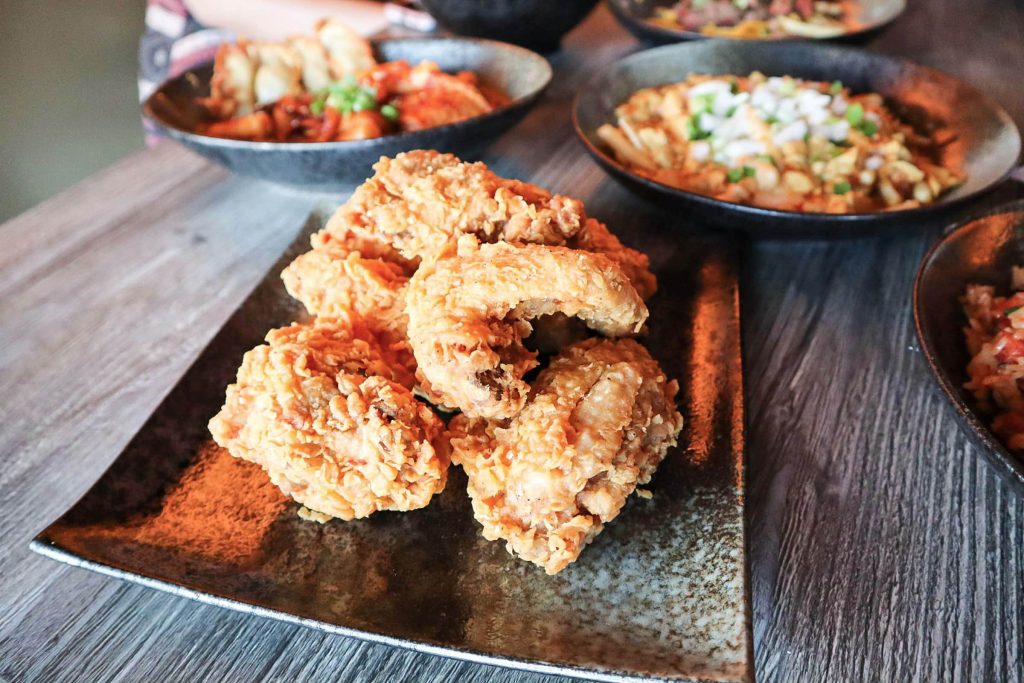 A rectangular plate with crispy fried chicken pieces sits on a wooden table, surrounded by bowls of various dishes, including vegetables and other side items.