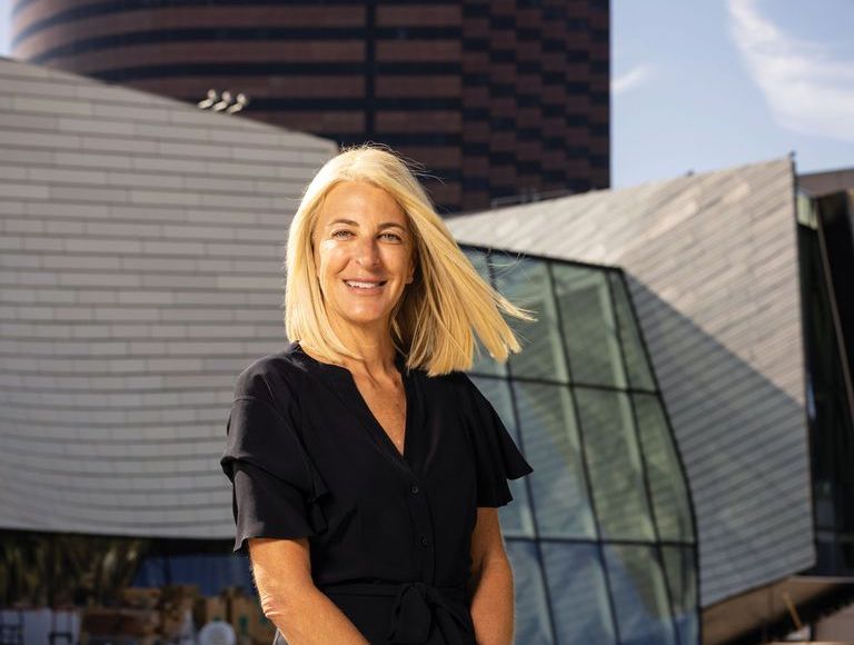 A smiling woman with blonde hair stands outdoors in front of modern, angular glass and metal buildings on a sunny day. She is wearing a short-sleeved black dress.