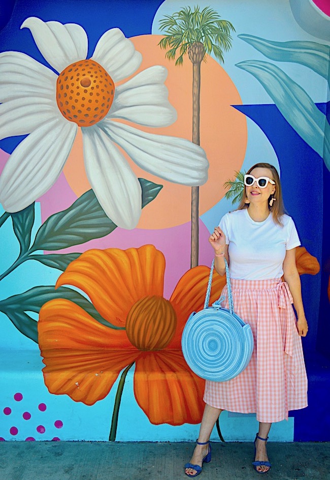 A woman in a white t-shirt, pink gingham skirt, blue sandals, and white sunglasses stands in front of a colorful mural with large flowers, leaves, and palm trees. She holds a round blue bag and looks to the side, smiling.