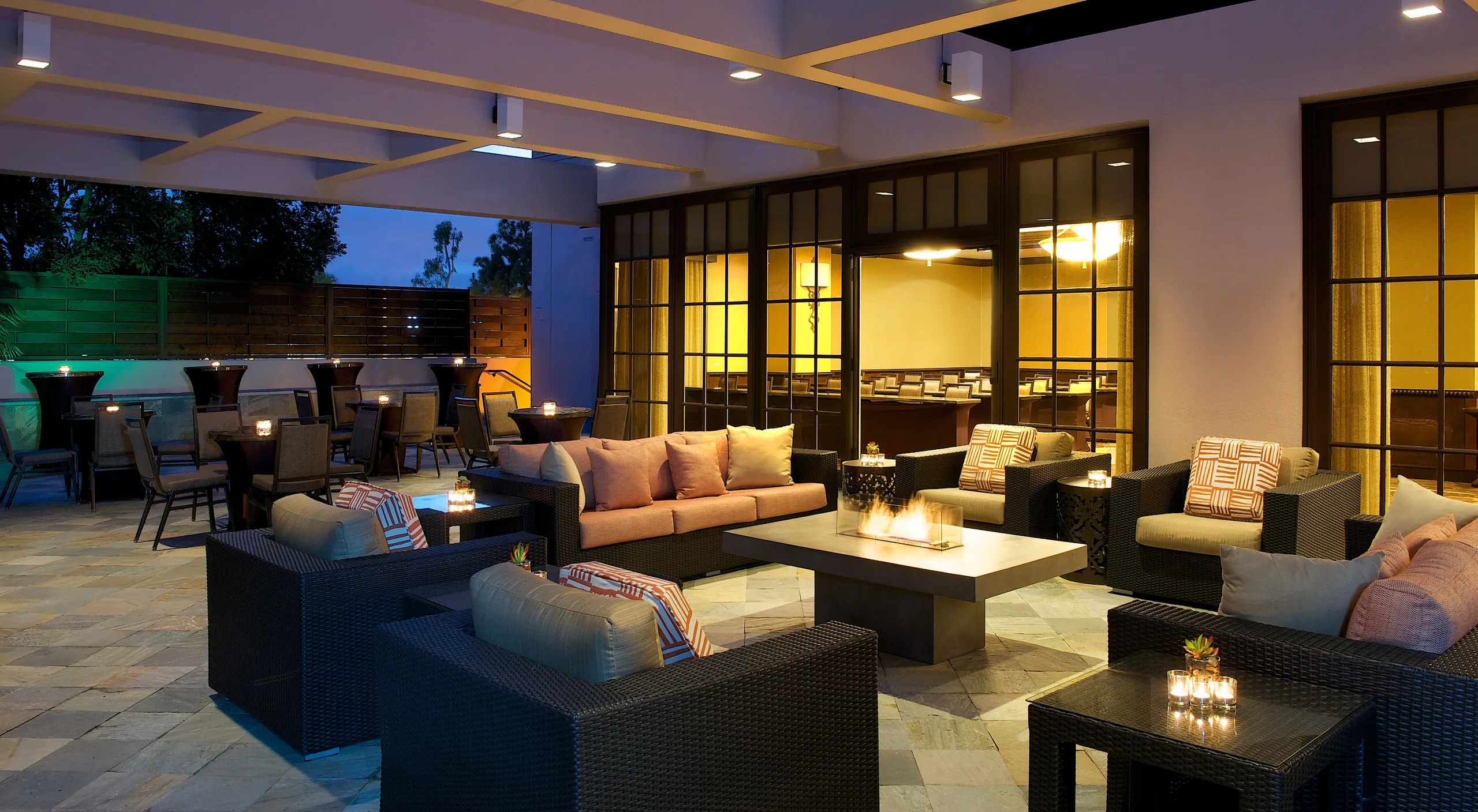 Outdoor patio lounge at Hilton Orange County Costa Mesa with cushioned wicker chairs, sofas, low tables, and lit candles on a stone floor. Warm lighting creates a cozy, inviting atmosphere near large windows and wooden partitions.