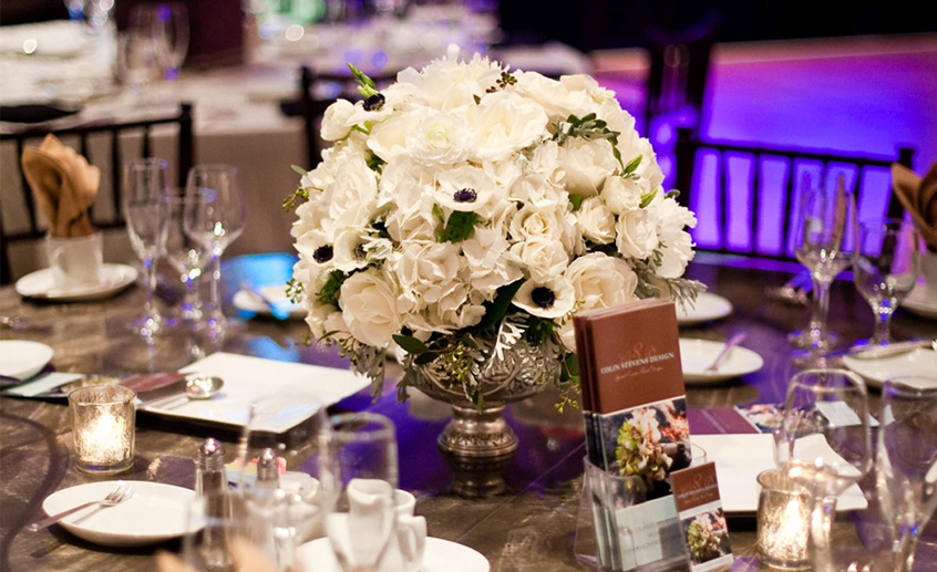 A round table set for an event at Hilton Orange County Costa Mesa features a large centerpiece of white flowers in a silver vase, surrounded by elegant place settings, menus, glassware, and candle votives against a softly lit backdrop.