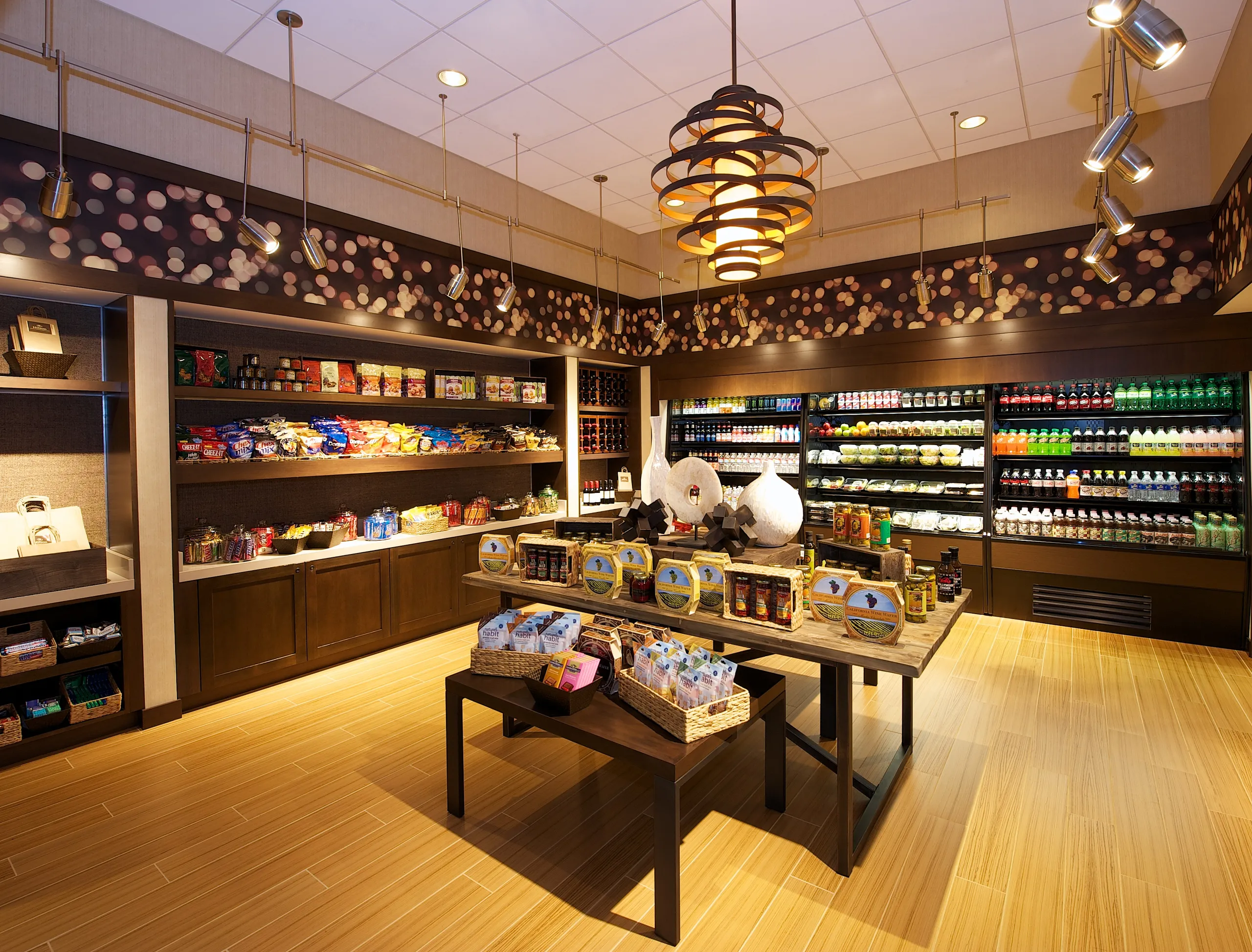 A well-lit, modern convenience store at Hilton Orange County Costa Mesa with shelves and coolers stocked with snacks, drinks, and packaged goods; a display table in the center features gift baskets and specialty items.