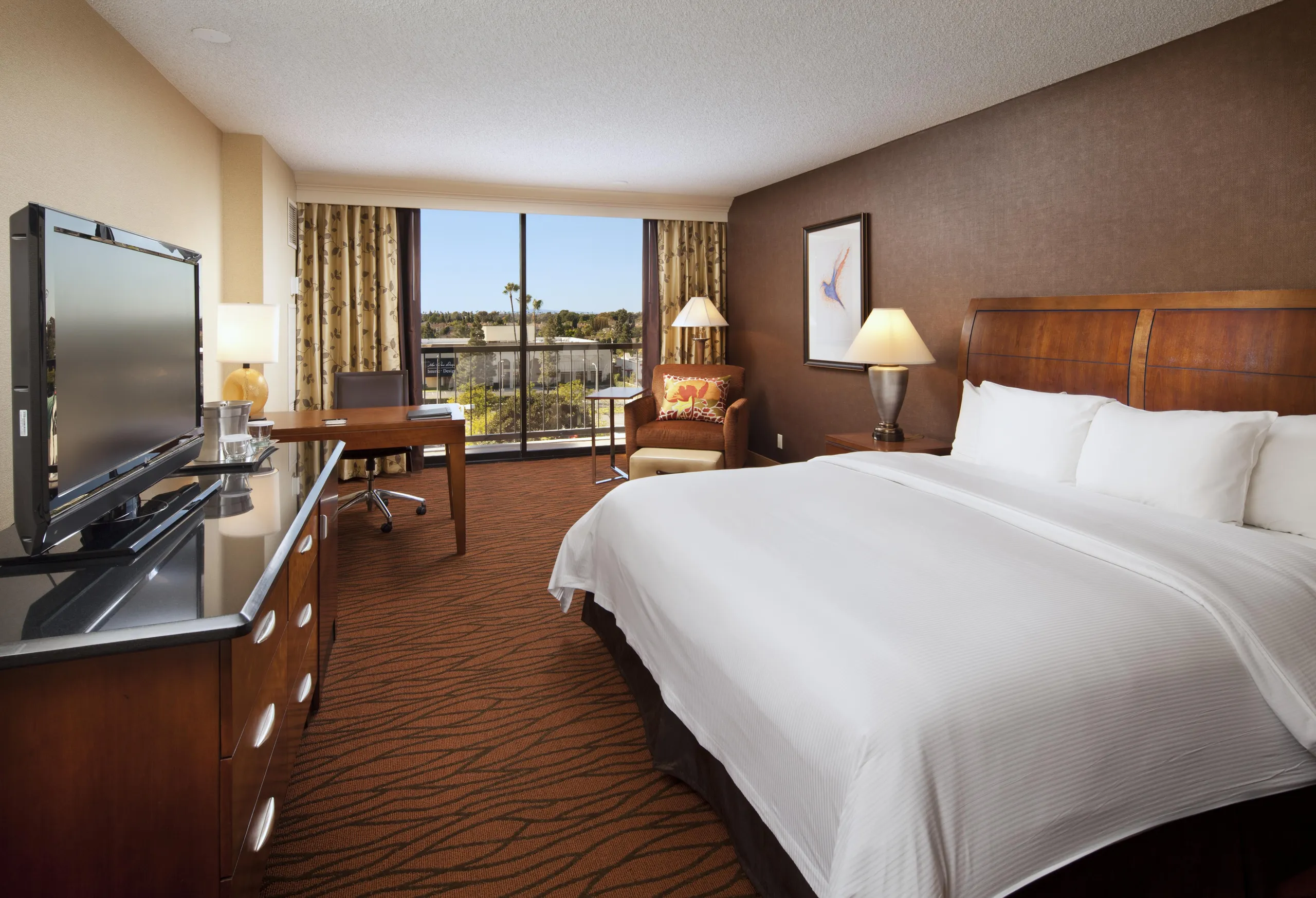 A modern Hilton Orange County Costa Mesa hotel room with a large bed, flat-screen TV on a dresser, desk with chair, armchair and lamp, plus large windows offering views of trees and city buildings outside.