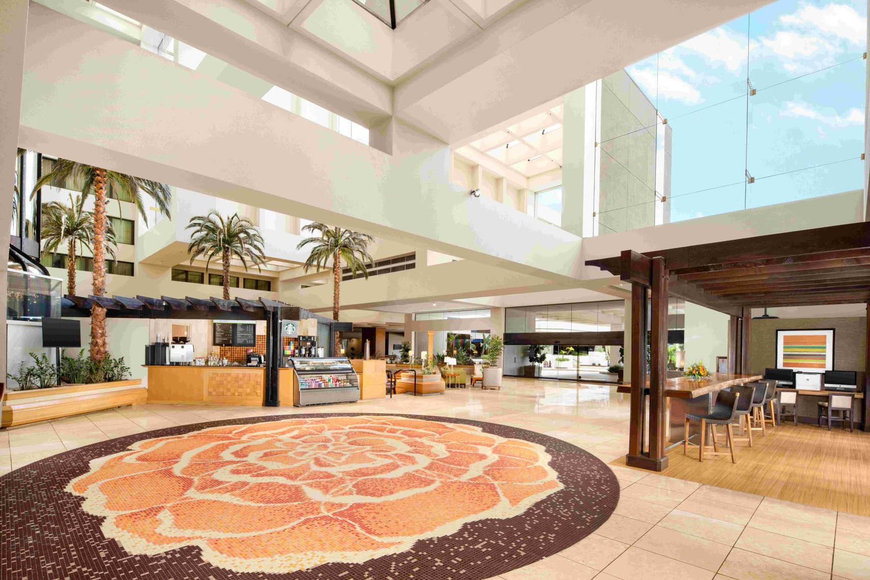 Spacious Hilton Orange County Costa Mesa hotel lobby with large windows, indoor palm trees, a café counter, modular seating, and a round orange floral-patterned rug on the tiled floor under natural light.