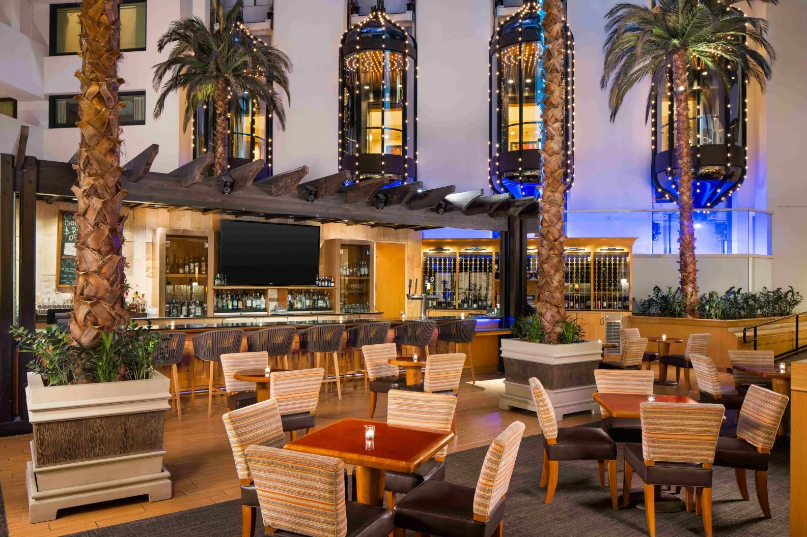 Modern hotel lobby bar at Hilton Orange County Costa Mesa, featuring wooden tables and striped chairs, surrounded by tall palm trees and illuminated glass elevators, creates a stylish and inviting atmosphere with warm and blue lighting.