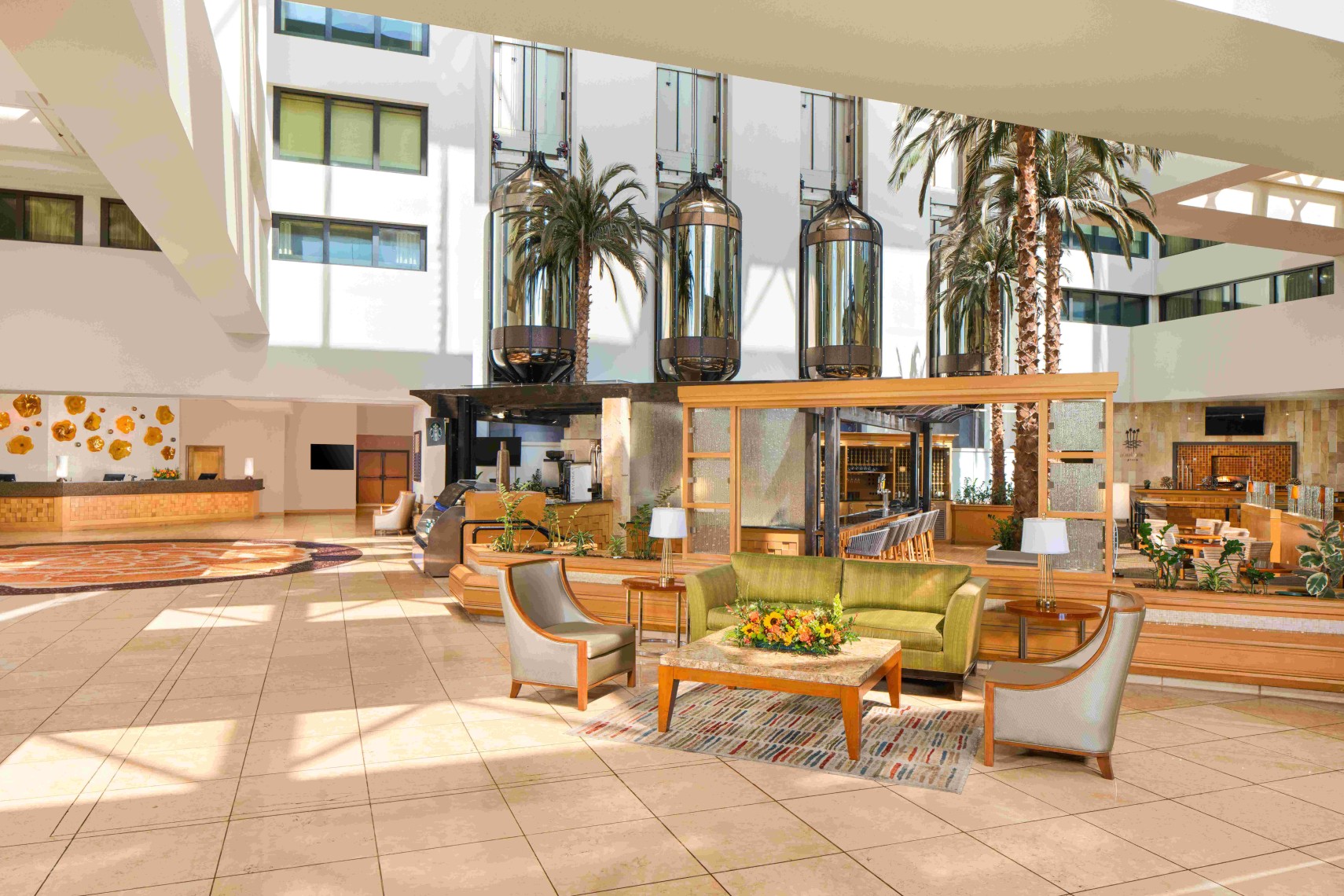 Spacious hotel lobby at Hilton Orange County Costa Mesa with modern decor, green sofa, armchairs, glass elevators, tall indoor palm trees, and abundant natural light from large windows and skylights. Reception desk visible in the background.